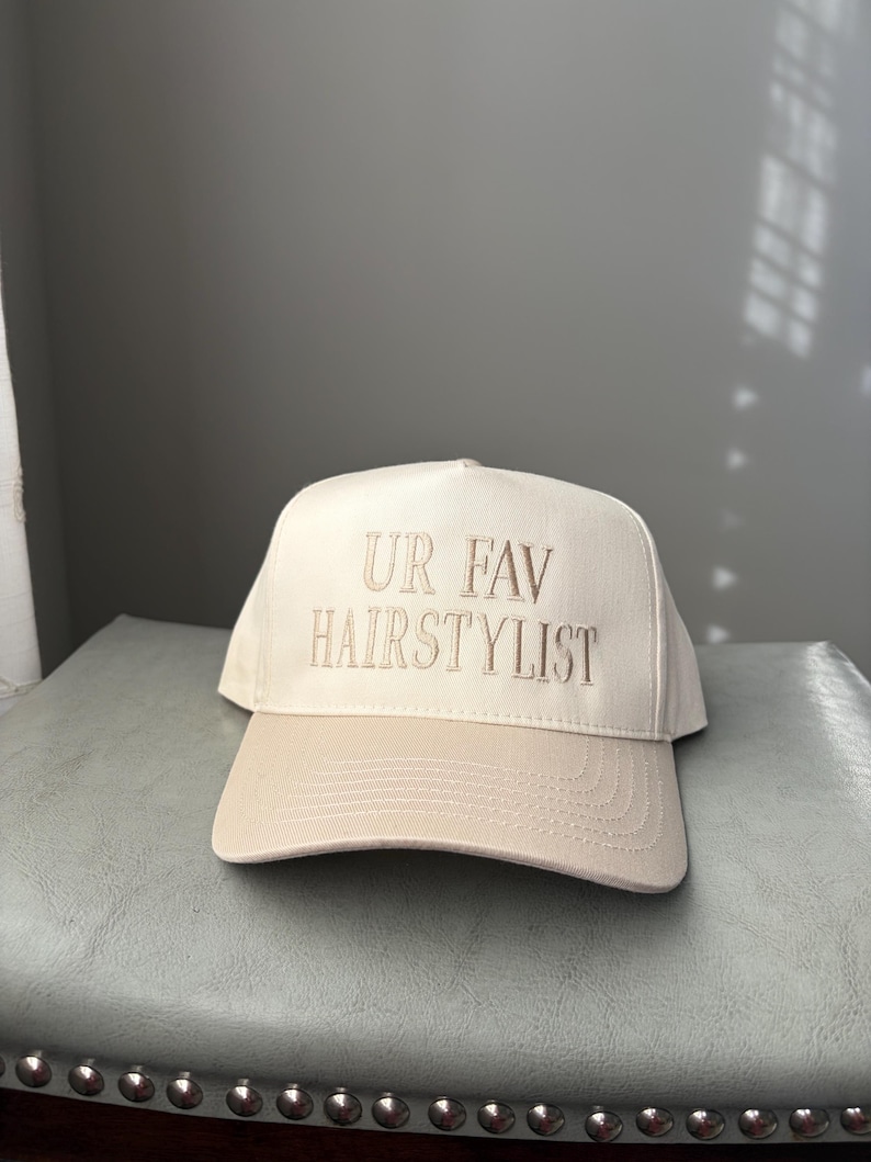 May include: A cream-coloured baseball cap with a tan brim. The phrase "UR FAV HAIRSTYLIST" is embroidered in a matching tan thread across the front of the cap. The cap is sitting on a light blue surface.