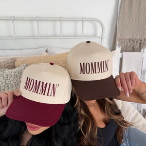 Puede incluir: Dos gorras de b&eacute;isbol con la palabra "Mommin'" impresa en la parte delantera. Una gorra tiene una corona color crema y una visera burdeos, mientras que la otra tiene una corona color crema y una visera marr&oacute;n. Ambas gorras tienen un bot&oacute;n marr&oacute;n oscuro en la parte superior.