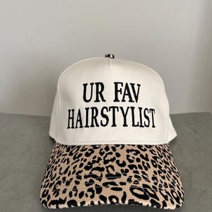 May include: A cream-coloured baseball cap with the embroidered text "UR FAV HAIRSTYLIST" in black. The peak has a tan and black leopard print. This cap is suitable for everyday wear.