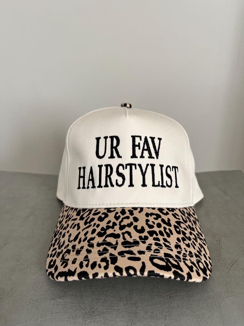May include: A cream-coloured baseball cap with the embroidered text "UR FAV HAIRSTYLIST" in black. The peak has a tan and black leopard print. This cap is suitable for everyday wear.