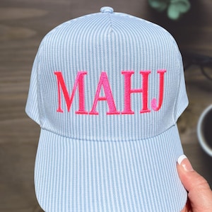 May include: A light blue and white striped baseball cap with the embroidered letters "MAHJ" in bright pink. The cap has a curved brim and a button on top. The website "shopsimplydelilah.com" is printed on the bottom.