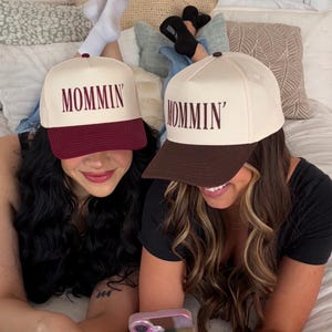 Puede incluir: Dos gorras de b&eacute;isbol con la palabra "MOMMIN'" bordada en la parte delantera. Una gorra tiene una corona color crema y una visera burdeos, mientras que la otra tiene una corona crema y una visera marr&oacute;n. Las gorras son usadas por dos personas.