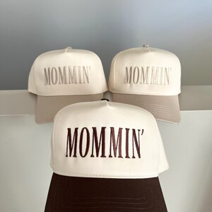 Puede incluir: Tres gorras de b&eacute;isbol con la palabra "MOMMIN'" bordada en la parte delantera. Las gorras tienen un panel frontal blanco y una visera marr&oacute;n. El texto est&aacute; bordado en marr&oacute;n a juego.