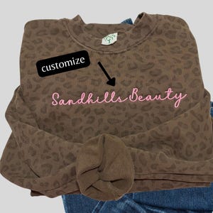 May include: A brown crewneck sweatshirt featuring an all-over dark brown leopard print pattern. The text "Sandhills Beauty" is embroidered in light pink script across the chest. A black rectangular tag with the word "customize" and an arrow.