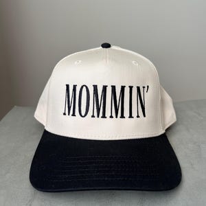 Puede incluir: Gorra de b&eacute;isbol con corona color crema y visera negra. La palabra "MOMMIN'" est&aacute; bordada en negro en la parte delantera. La gorra tiene un bot&oacute;n negro en la parte superior.