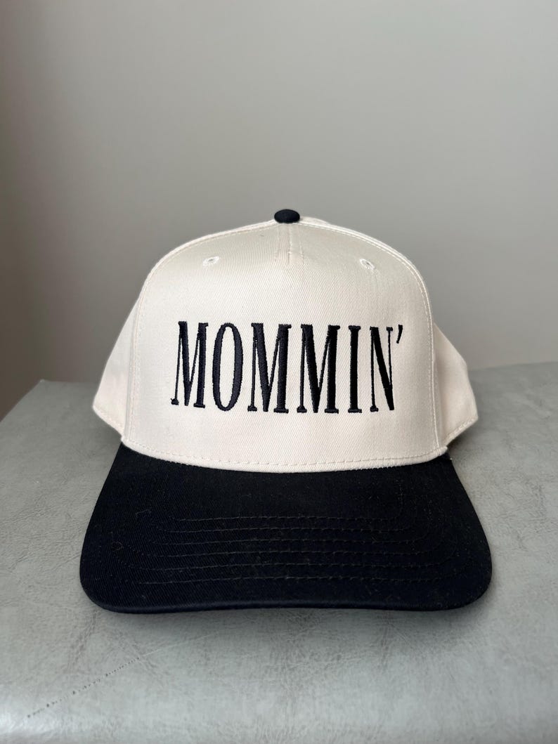 Puede incluir: Gorra de b&eacute;isbol con corona color crema y visera negra. La palabra "MOMMIN'" est&aacute; bordada en negro en la parte delantera. La gorra tiene un bot&oacute;n negro en la parte superior.