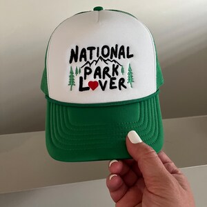 May include: A white and green trucker hat with the text "NATIONAL PARK LOVER" embroidered in black with a red heart. The hat has a green mesh back.