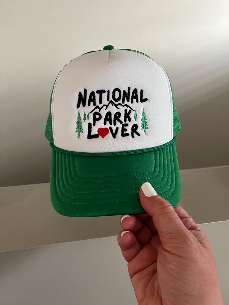 May include: A white and green trucker hat with the text "NATIONAL PARK LOVER" embroidered in black with a red heart. The hat has a green mesh back.