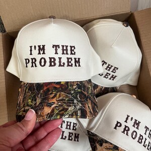 May include: A set of four white baseball caps with a camouflage brim. The caps have brown embroidery that reads "I'm the Problem".