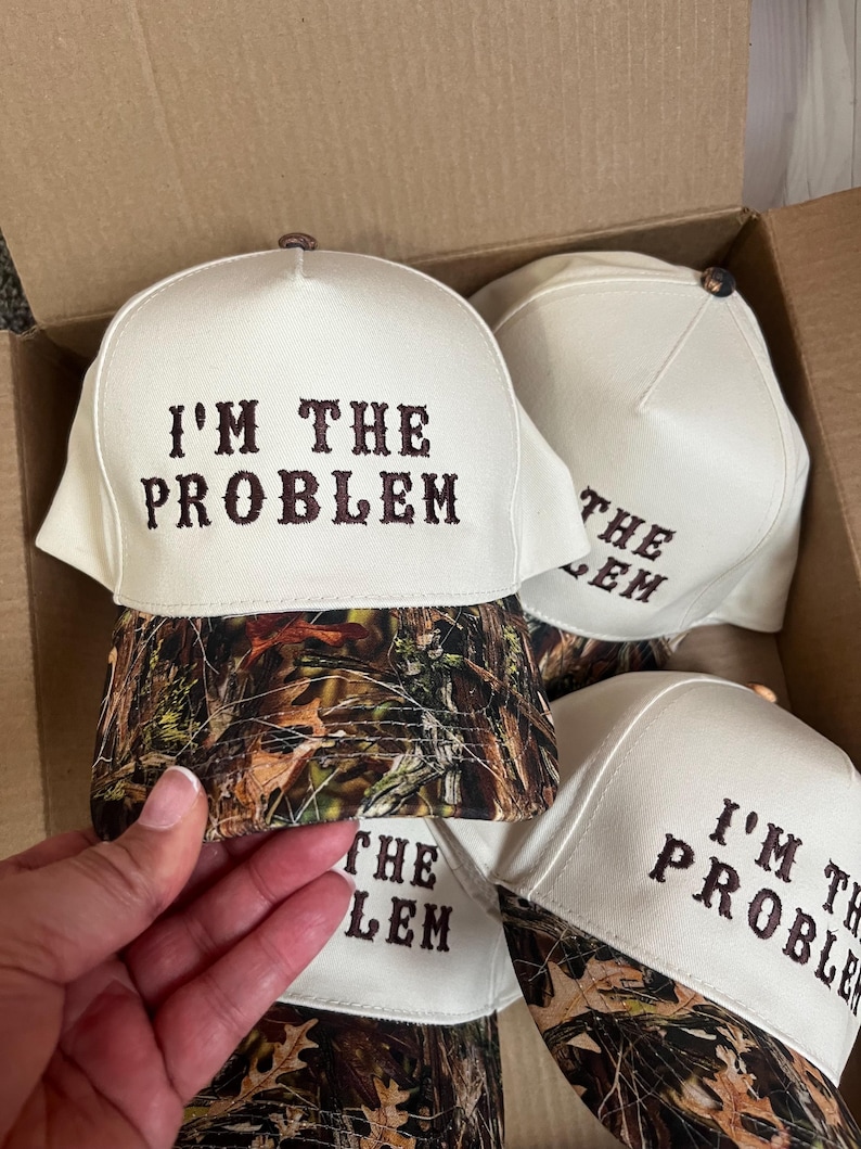 May include: A set of four white baseball caps with a camouflage brim. The caps have brown embroidery that reads "I'm the Problem".