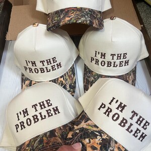 May include: Four white baseball caps with a camouflage brim. Each cap has the text "I'M THE PROBLEM" embroidered in brown on the front.