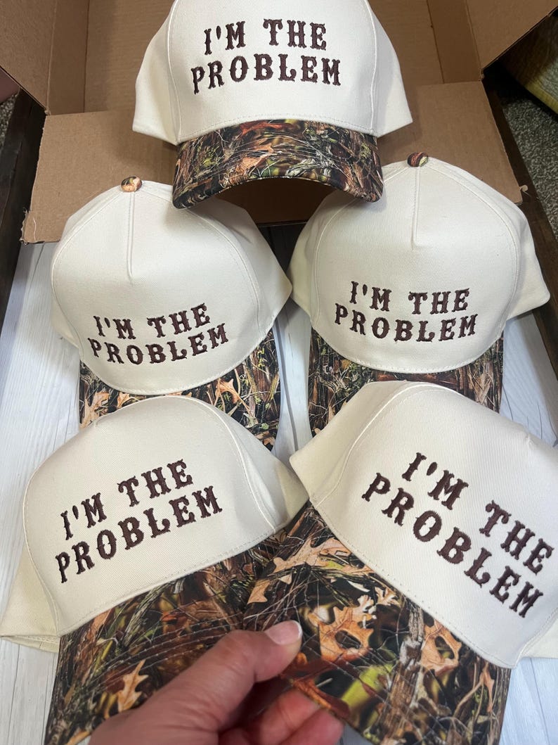 May include: Four white baseball caps with a camouflage brim. Each cap has the text "I'M THE PROBLEM" embroidered in brown on the front.