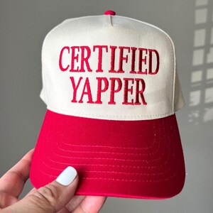 May include: A white and red trucker hat with the embroidered text "CERTIFIED YAPPER" in red. The hat features a red brim and a white front panel with the text. The top button is red.
