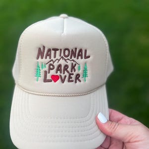 May include: A beige trucker hat with a brown embroidered design that says "National Park Lover" with a red heart and green pine trees.