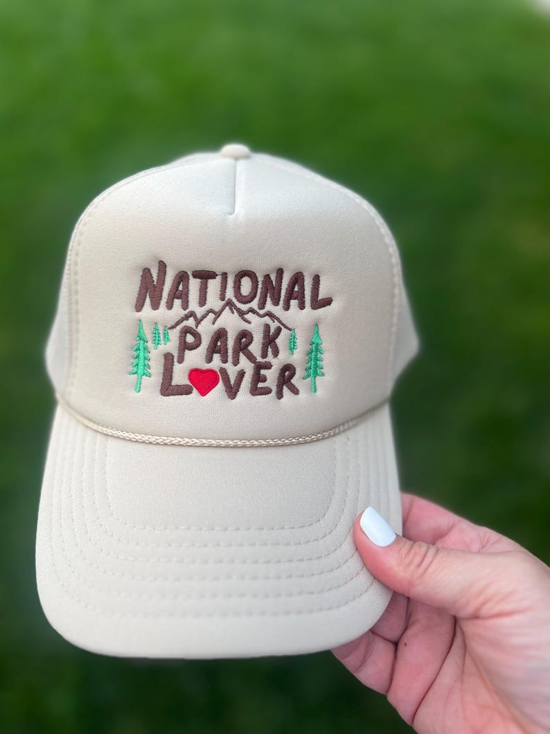 May include: A beige trucker hat with a brown embroidered design that says "National Park Lover" with a red heart and green pine trees.