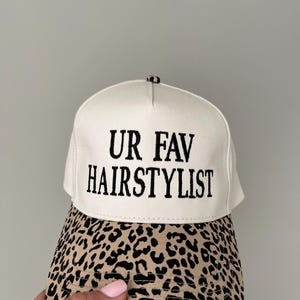 May include: A cream-coloured baseball cap with the text "UR FAV HAIRSTYLIST" in black. The peak has a tan and black leopard print design. A fashionable headwear item.