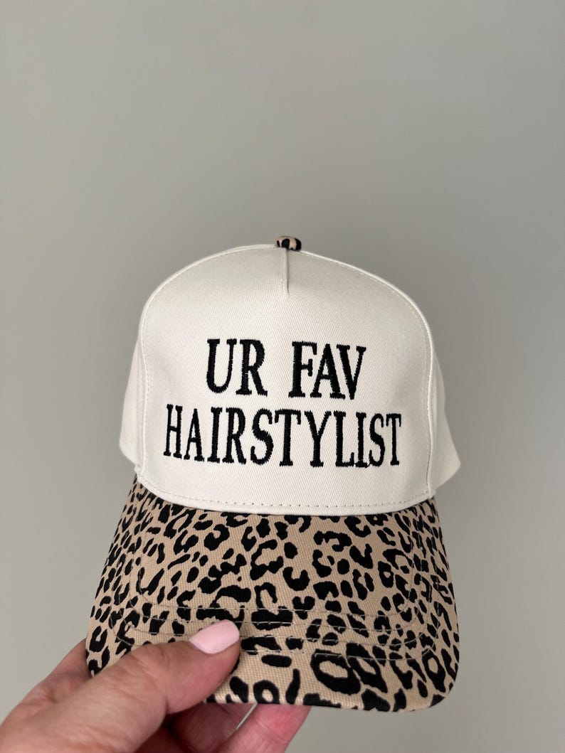 May include: A cream-coloured baseball cap with the text "UR FAV HAIRSTYLIST" in black. The peak has a tan and black leopard print design. A fashionable headwear item.