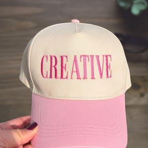 May include: A baseball cap with a cream-colored crown and a pink bill. The word "CREATIVE" is embroidered in pink across the front. The cap has a pink button on top.