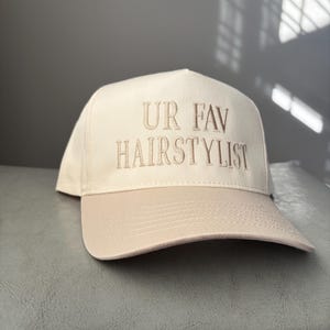 May include: A beige baseball cap with the embroidered text "UR FAV HAIRSTYLIST" in a matching beige thread. The cap has a curved brim and a structured crown, suitable for hairstylists.