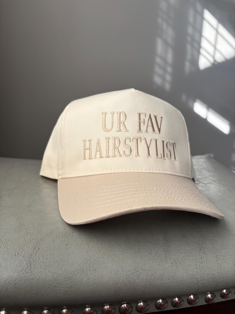 May include: A beige baseball cap with the embroidered text "UR FAV HAIRSTYLIST" in a matching beige thread. The cap has a curved brim and a structured crown, suitable for hairstylists.