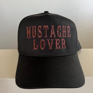 Pode incluir: Um bon&eacute; de beisebol preto com a frase "MUSTACHE LOVER" bordada em vermelho na frente.