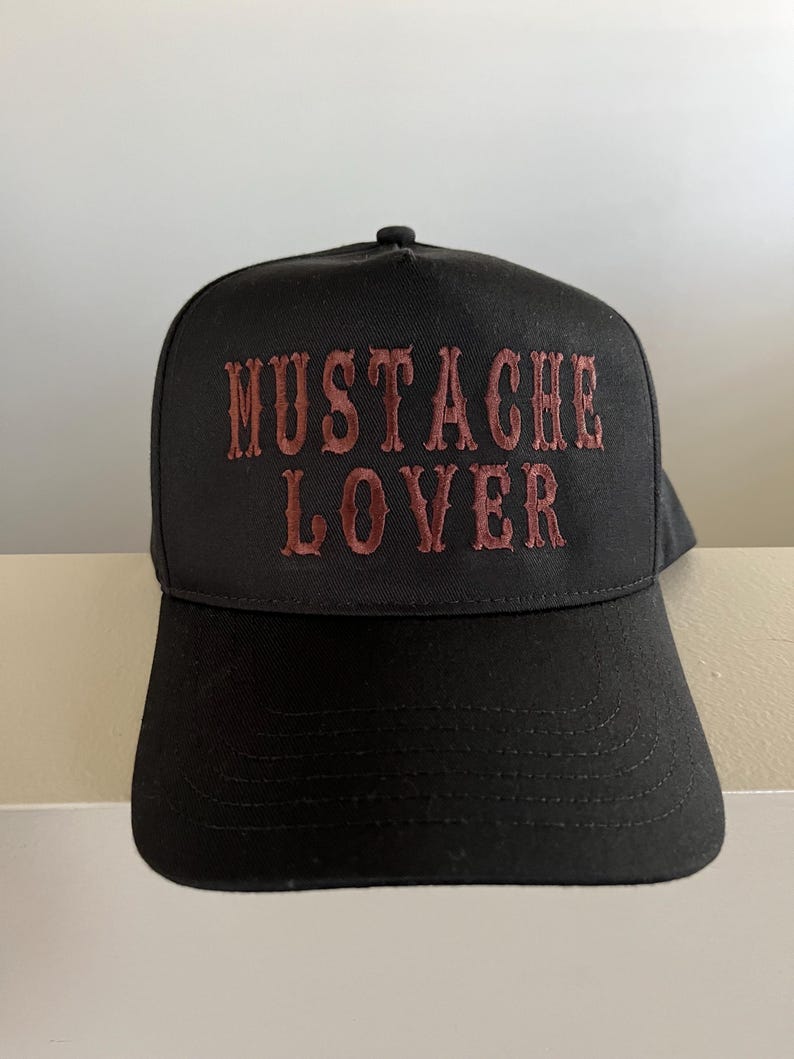Pode incluir: Um bon&eacute; de beisebol preto com a frase "MUSTACHE LOVER" bordada em vermelho na frente.