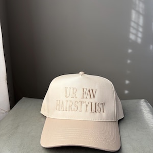 May include: A beige baseball cap with a curved peak. The cap has the embroidered text "UR FAV HAIRSTYLIST" in a matching beige thread. The hat is made of a soft fabric and has a button on top.