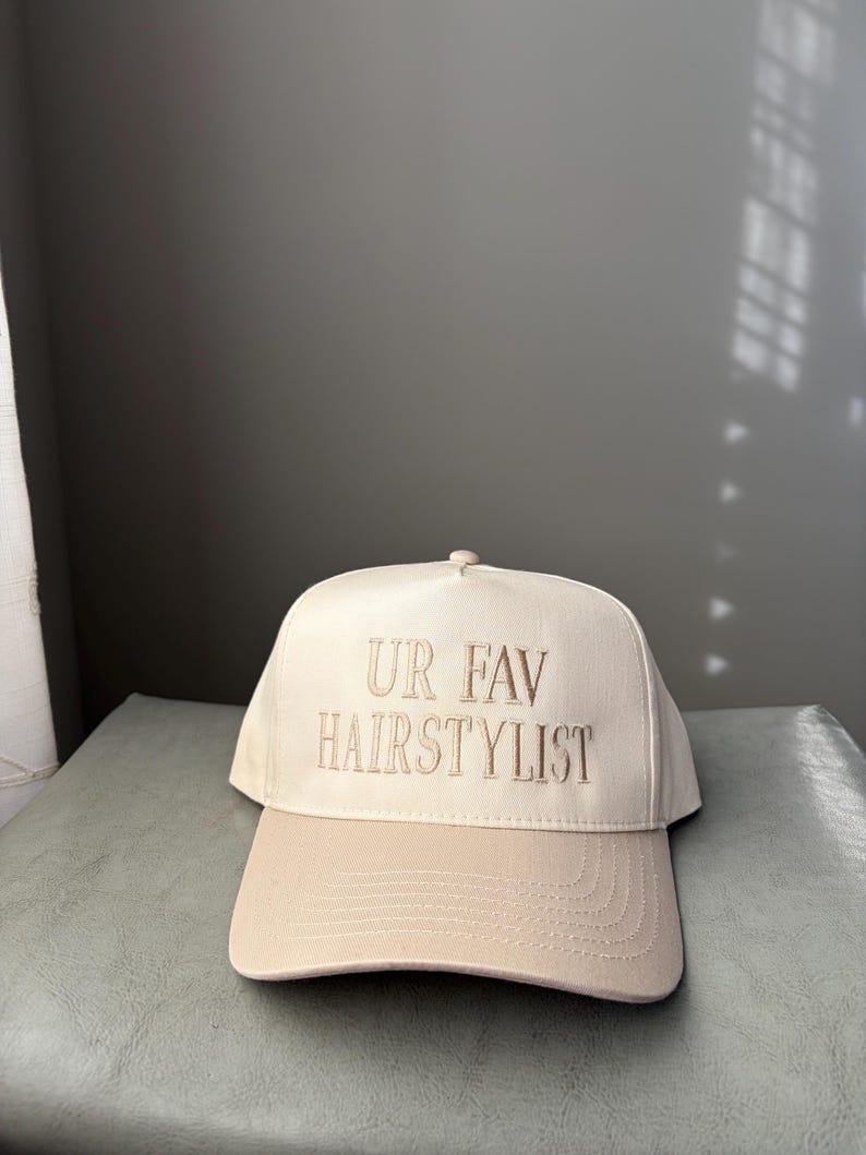 May include: A beige baseball cap with a curved peak. The cap has the embroidered text "UR FAV HAIRSTYLIST" in a matching beige thread. The hat is made of a soft fabric and has a button on top.