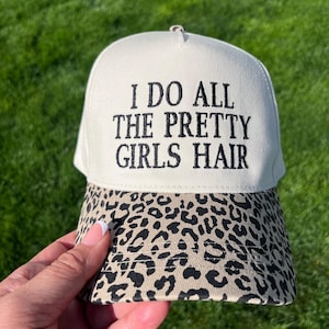 May include: A white baseball cap with a black leopard print brim. The front of the cap has black embroidery that reads "I do all the pretty girls hair."