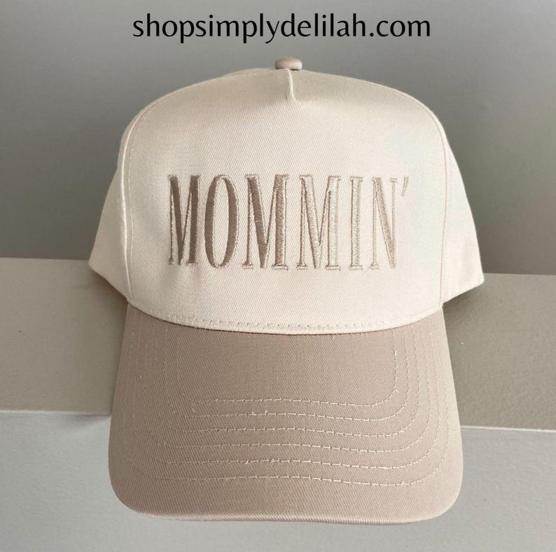 Puede incluir: Gorra de b&eacute;isbol beige con visera en contraste marr&oacute;n claro. La palabra "MOMMIN'" est&aacute; bordada con hilo marr&oacute;n claro a juego en la parte delantera. El sitio web shopsimplydelilah.com est&aacute; impreso en la parte superior.