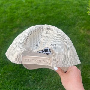 May include: A beige mesh trucker hat with a black and white animal print patch on the front. The hat has a black adjustable strap with a plastic buckle.