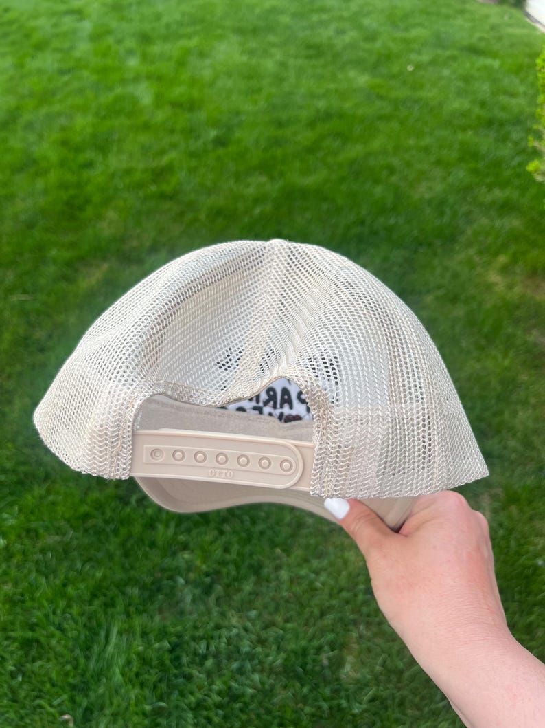 May include: A beige mesh trucker hat with a black and white animal print patch on the front. The hat has a black adjustable strap with a plastic buckle.