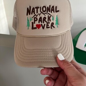 May include: A beige trucker hat with a brown embroidered design that says "National Park Lover" with a red heart. The hat has a mesh back.