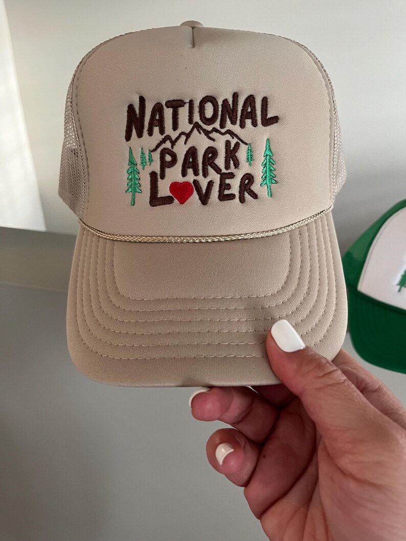 May include: A beige trucker hat with a brown embroidered design that says "National Park Lover" with a red heart. The hat has a mesh back.