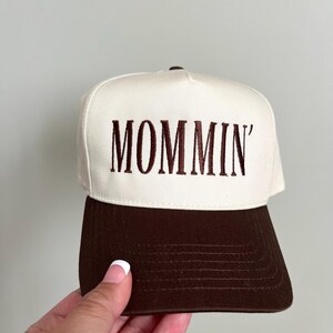 Puede incluir: Gorra de b&eacute;isbol con corona blanca y visera marr&oacute;n. La palabra "MOMMIN'" est&aacute; bordada en marr&oacute;n en la parte delantera. La gorra tiene un dise&ntilde;o cl&aacute;sico de seis paneles.