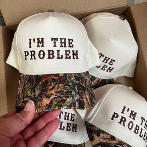 May include: Four white baseball caps with a camouflage brim. The caps have brown embroidery that reads "I'm the Problem" and "The Problem".