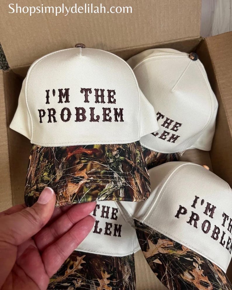 May include: Four white baseball caps with a camouflage brim. The caps have brown embroidery that reads "I'm the Problem" and "The Problem".