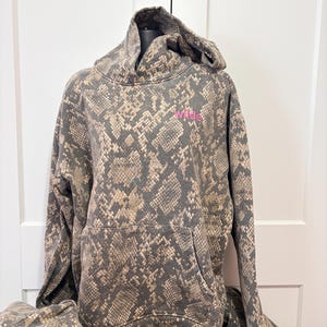 May include: A gray and brown snakeskin-patterned hoodie with a kangaroo pocket and a drawstring hood. The word "wilde" is printed in pink on the chest. The hoodie is displayed on a mannequin. A matching pair of pants is visible.