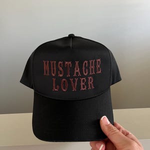 Pode incluir: Bon&eacute; de beisebol preto com uma frase bordada em vermelho "MUSTACHE LOVER" na frente.