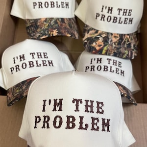 May include: Five white baseball caps with a camouflage brim. Each cap has the text "I'M THE PROBLEM" embroidered in brown on the front.