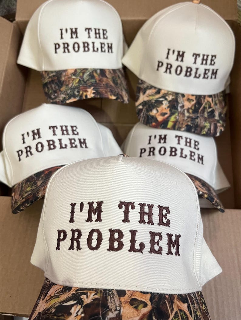 May include: Five white baseball caps with a camouflage brim. Each cap has the text "I'M THE PROBLEM" embroidered in brown on the front.