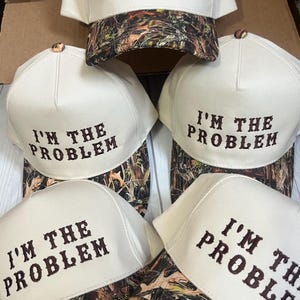 May include: Five white baseball caps with a camouflage pattern on the underside. Each cap has the phrase "I'm the Problem" embroidered in brown on the front.