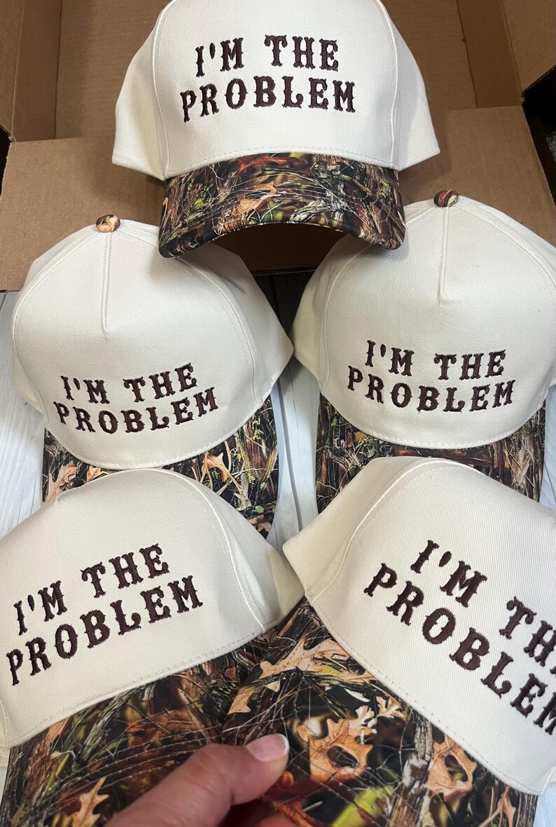 May include: Five white baseball caps with a camouflage pattern on the underside. Each cap has the phrase "I'm the Problem" embroidered in brown on the front.