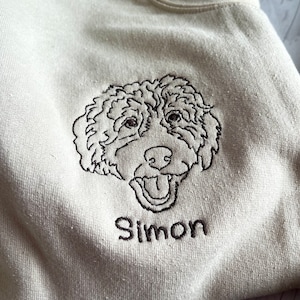 May include: A beige sweatshirt with an embroidered image of a dog's head and the name "Simon" underneath.