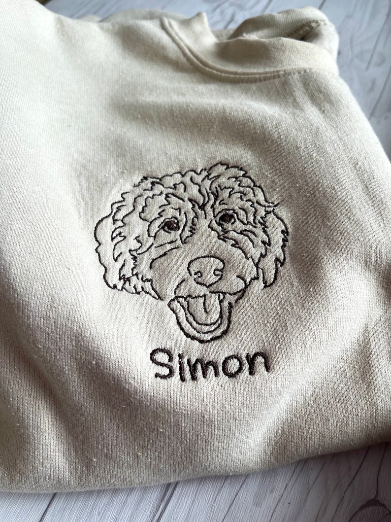 May include: A beige sweatshirt with an embroidered image of a dog's head and the name "Simon" underneath.