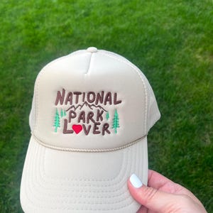 May include: A beige trucker hat with a brown embroidered design that says "National Park Lover" with a red heart. The hat has a mesh back.