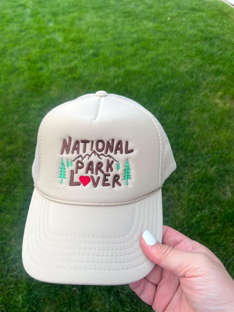 May include: A beige trucker hat with a brown embroidered design that says "National Park Lover" with a red heart. The hat has a mesh back.