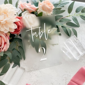 May include: A frosted acrylic table number with the text "Table 2" in white calligraphy. The number is displayed on a white surface with pink ribbon and greenery.