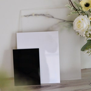 May include: Three blank acrylic sheets of different sizes, one black, one white, and one frosted white. The sheets are stacked on a wooden surface.