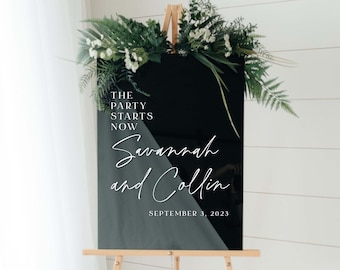 May include: Black acrylic wedding sign with white text that reads "The party starts now Savannah and Collin September 3, 2023". The sign is decorated with greenery and white flowers. Shown size is 18 inches by 24 inches.